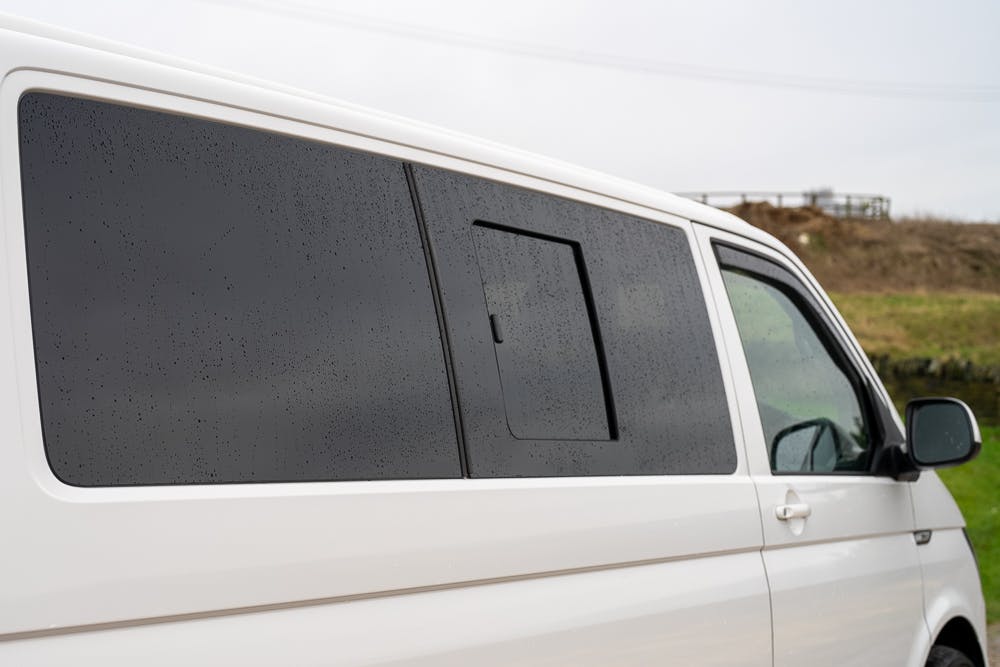 A 2019 Volkswagen Transporter T28 Trendline TDI is parked outdoors. The side windows, which are tinted, have water droplets on them, indicating recent rain. The rear side window has a smaller rectangular window section that can be opened, currently closed.