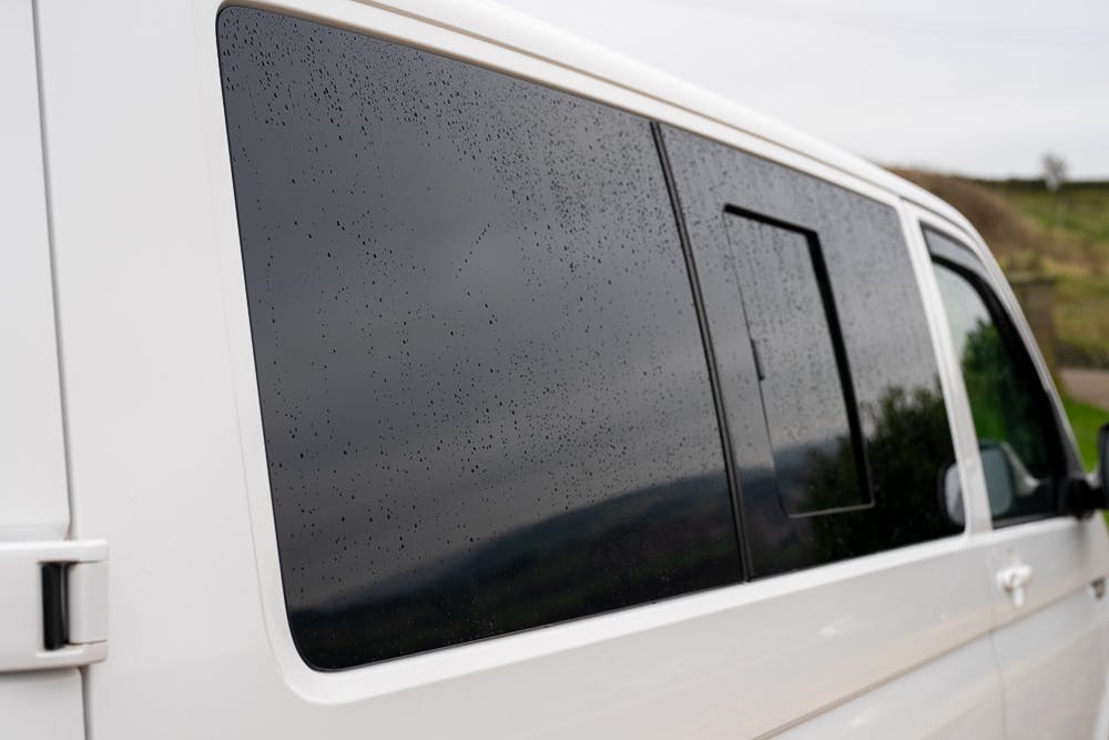 A 2019 Volkswagen Transporter T28 Trendline TDI with dark-tinted windows is parked outdoors. The exterior of the white vehicle is covered in water droplets, indicating recent rainfall. In the slightly blurred background, a mix of greenery and overcast skies can be seen.
