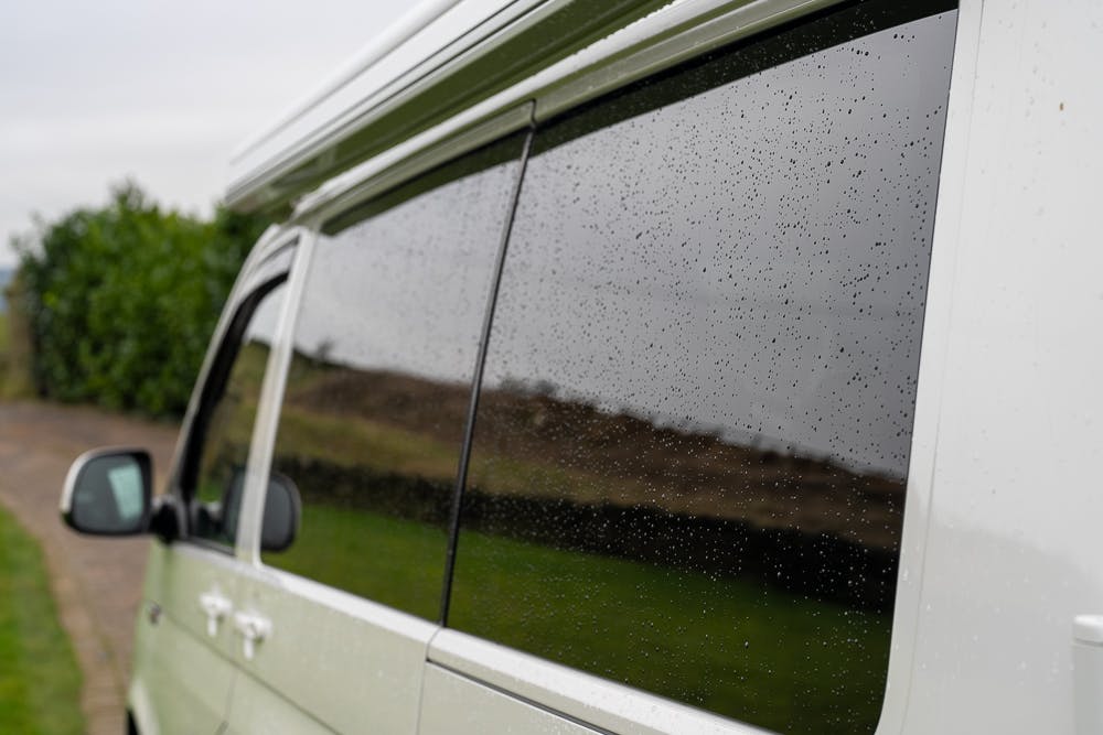 A close-up of the side of a 2019 Volkswagen Transporter T28 Trendline TDI showcasing its dark-tinted windows with raindrops on the glass. The background shows a blurred view of a grassy field and green bushes along a paved pathway. The sky appears overcast.
