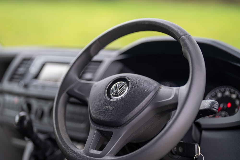 A close-up view of the steering wheel and dashboard of a 2019 Volkswagen Transporter T28 Trendline TDI. The Volkswagen logo is visible on the center of the steering wheel. The car’s interior includes a digital display and various control buttons, with blurred greenery in the background.