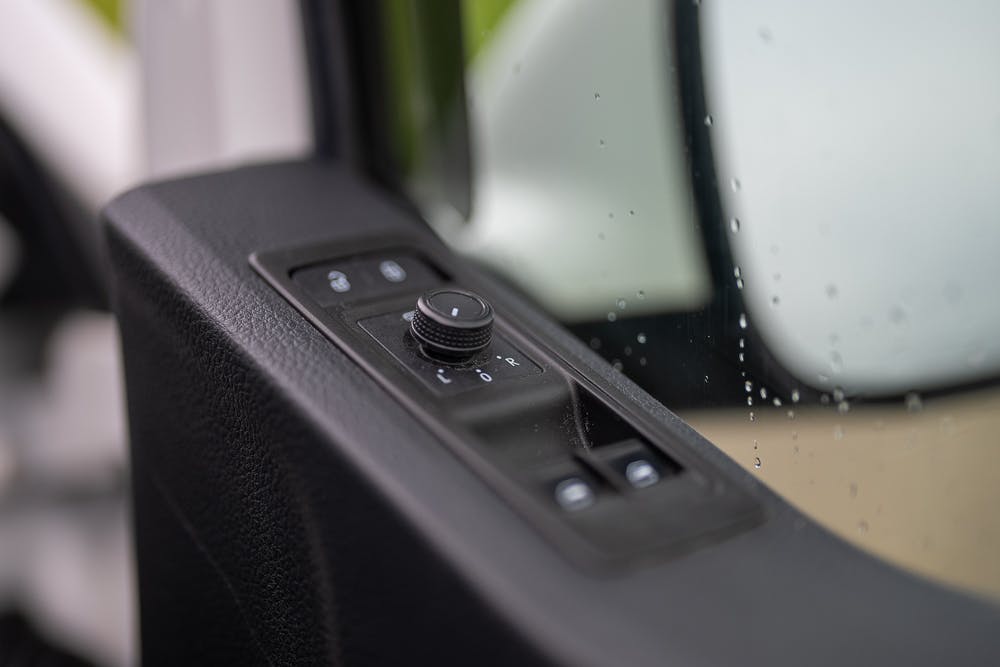 Close-up view of a 2019 Volkswagen Transporter T28 Trendline TDI's side mirror control panel on the driver's door. The panel includes a rotary knob for adjusting the mirror position and buttons for different settings. The panel is next to the side window, which has water droplets on it.