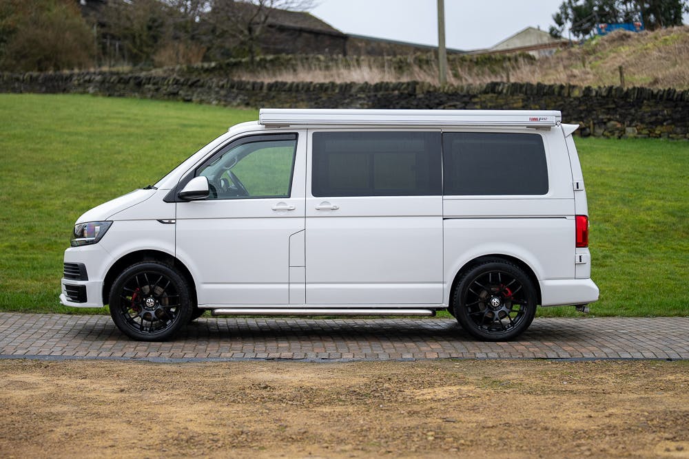 A 2019 Volkswagen Transporter T28 Trendline TDI with dark-tinted windows is parked on a paved surface, adorned by grass and a stone wall in the background. The van, featuring black alloy wheels, appears to be a modified camper van.
