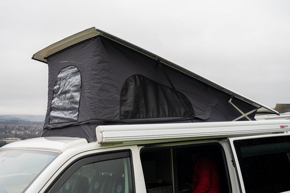 A white 2019 Volkswagen Transporter T28 Trendline TDI is equipped with a pop-up roof tent that is extended. The tent, dark gray with mesh windows, shelters a partially visible person inside the vehicle. The sky is overcast, and some landscape can be seen in the background.
