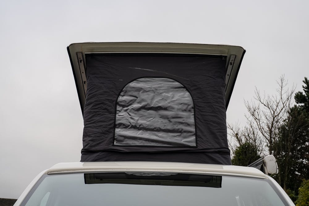 Close-up of a 2019 Volkswagen Transporter T28 Trendline TDI with an elevated black pop-up tent on the roof. The tent, featuring a rounded door flap, is extended vertically, suggesting it’s in camping mode. The background contains overcast sky and tree branches.