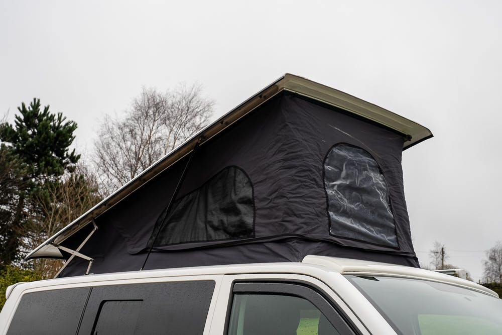 A 2019 Volkswagen Transporter T28 Trendline TDI features an elevated rooftop tent. The tent is black with clear plastic windows and appears to be set up for camping. In the background, trees stand under overcast skies.