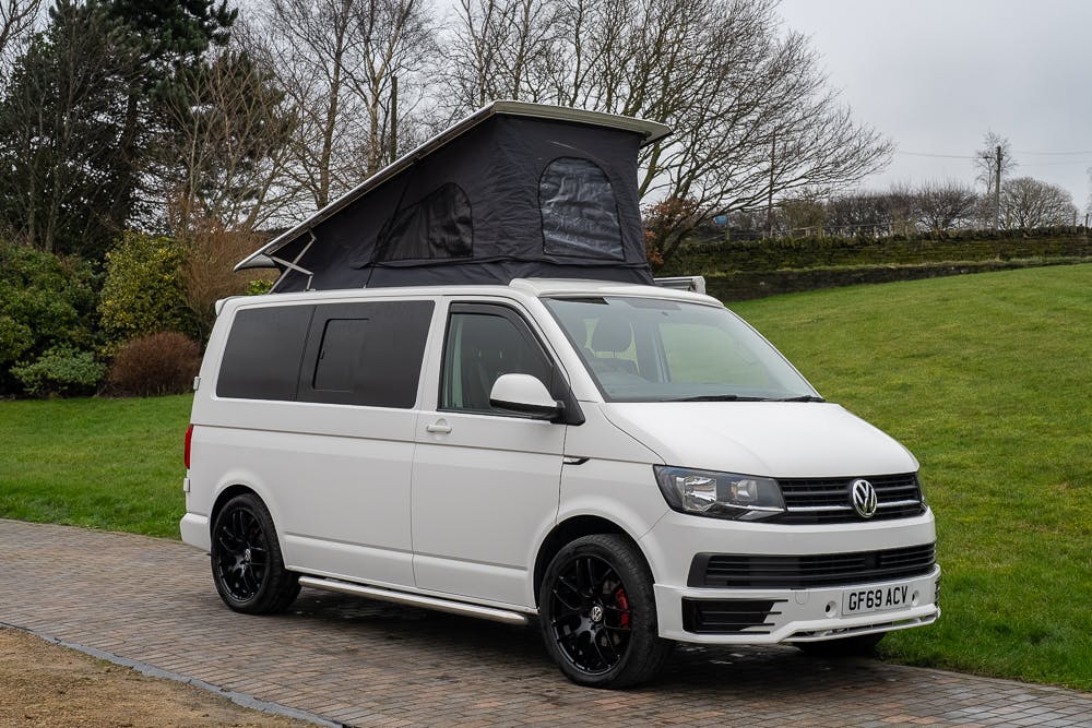 A white 2019 Volkswagen Transporter T28 Trendline TDI camper van is parked on a paved surface. The pop-top roof is elevated, creating additional space above. The surrounding area includes grass and trees, with a cloudy sky in the background. The license plate reads "GF69 ACV.