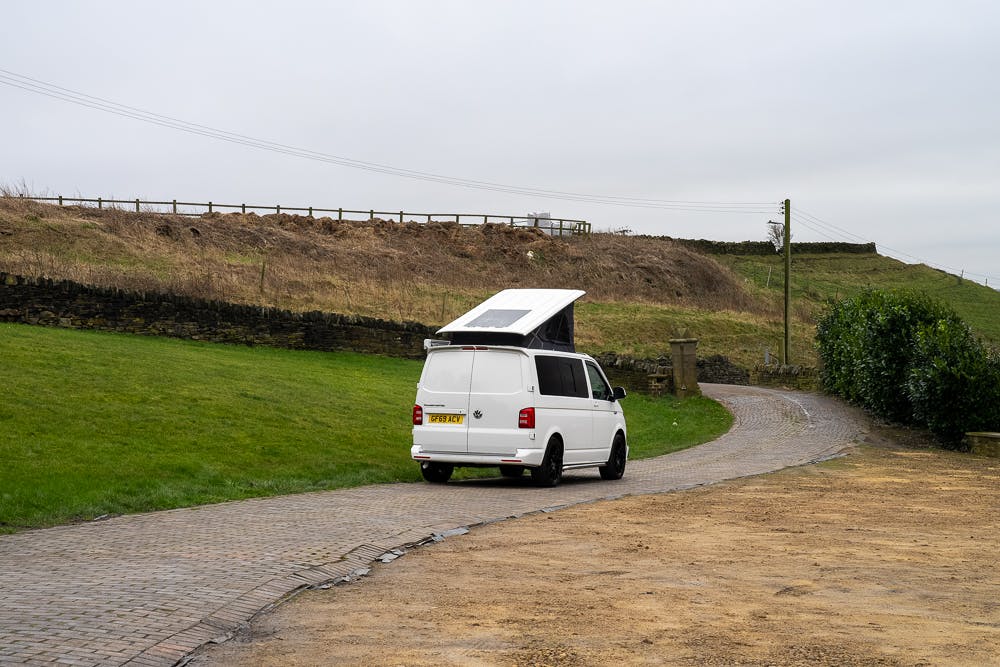 A white 2019 Volkswagen Transporter T28 Trendline TDI with a raised roof is parked on a path bordered by a grassy area and a stone wall. The sky is overcast, and the background includes a hill with a wooden fence.