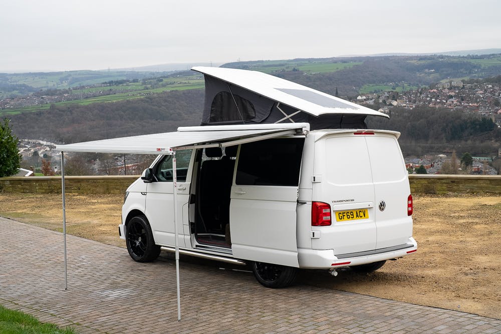 A white 2019 Volkswagen Transporter T28 Trendline TDI camper van with a UK license plate is parked beside a paved area, pop-up roof extended and side awning deployed. The van's side door is open, revealing the interior. In the background, there's a view of a rural town with hills and fields.