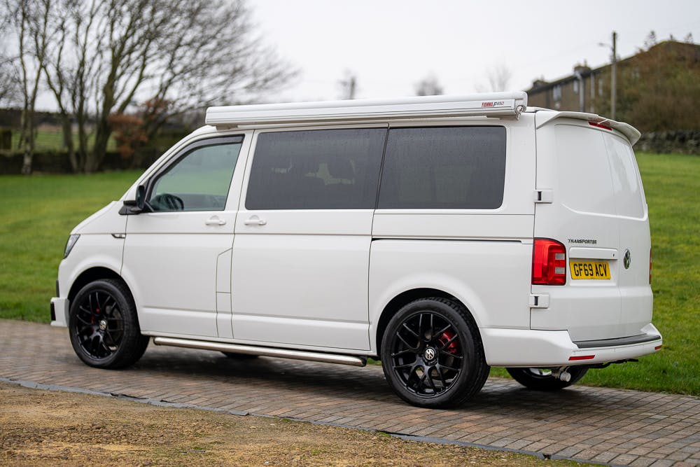 A white 2019 Volkswagen Transporter T28 Trendline TDI with black wheels is parked on a stone-paved area. The van, featuring dark-tinted windows and a retractable awning on the side, is surrounded by grass and trees. Its license plate reads "GF59 LXT.