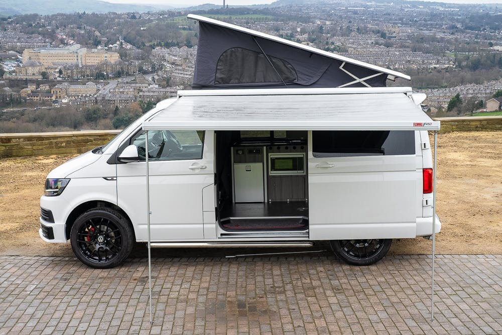 A 2019 Volkswagen Transporter T28 Trendline TDI camper van with a raised roof and extended awning is parked on a paved area. The side door is open, revealing the interior, which includes a kitchen unit. The background features a scenic view of a town and surrounding landscape.
