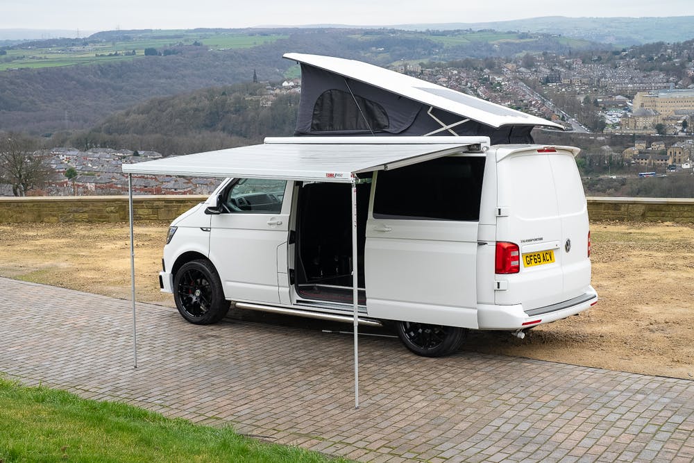 A 2019 Volkswagen Transporter T28 Trendline TDI with the registration plate "GF69 KXW" is parked on a paved surface with an extended awning and pop-up roof. The van's side door is open, revealing the interior. It is set against a backdrop of a hilly landscape and a town in the distance.