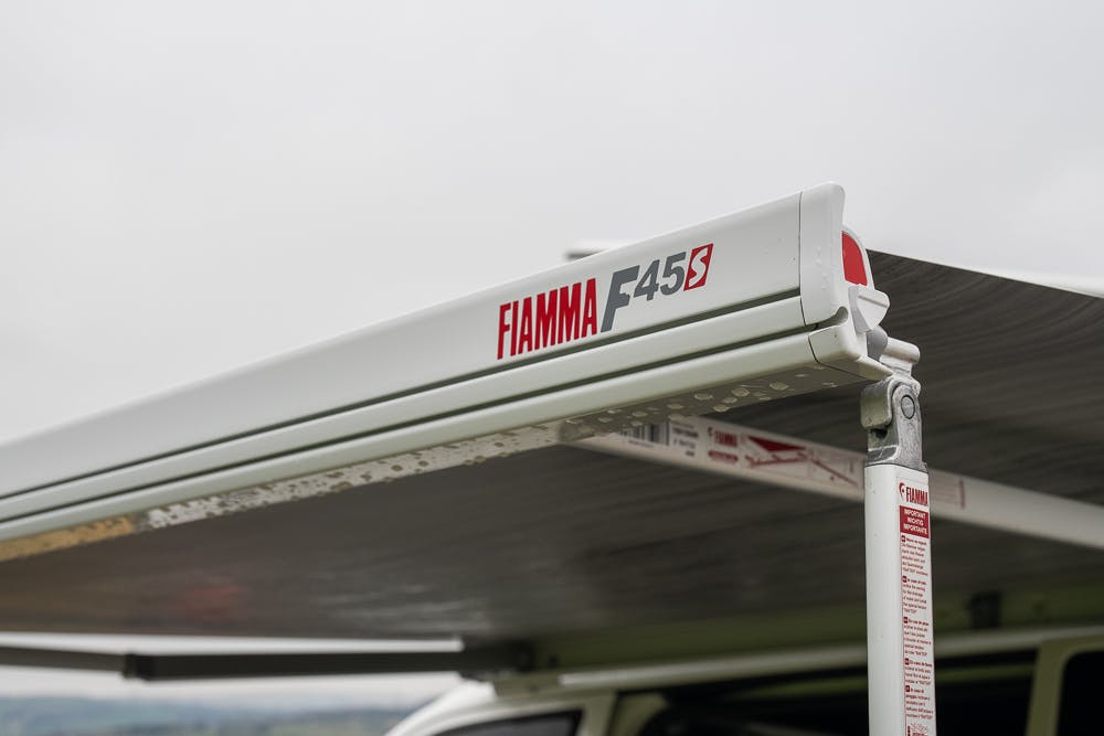 A close-up image of the Fiamma F45S awning attached to a 2019 Volkswagen Transporter T28 Trendline TDI. The extending awning is partially open, providing shade. The branding and model information of the awning are visible on the casing. The background shows an overcast sky and open land.