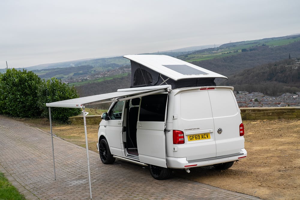 A 2019 Volkswagen Transporter T28 Trendline TDI with a raised roof and extended awning is parked on a brick-paved area overlooking a hilly landscape. The vehicle's license plate reads "GF69 ACV." The van's side door is open, revealing part of the interior.