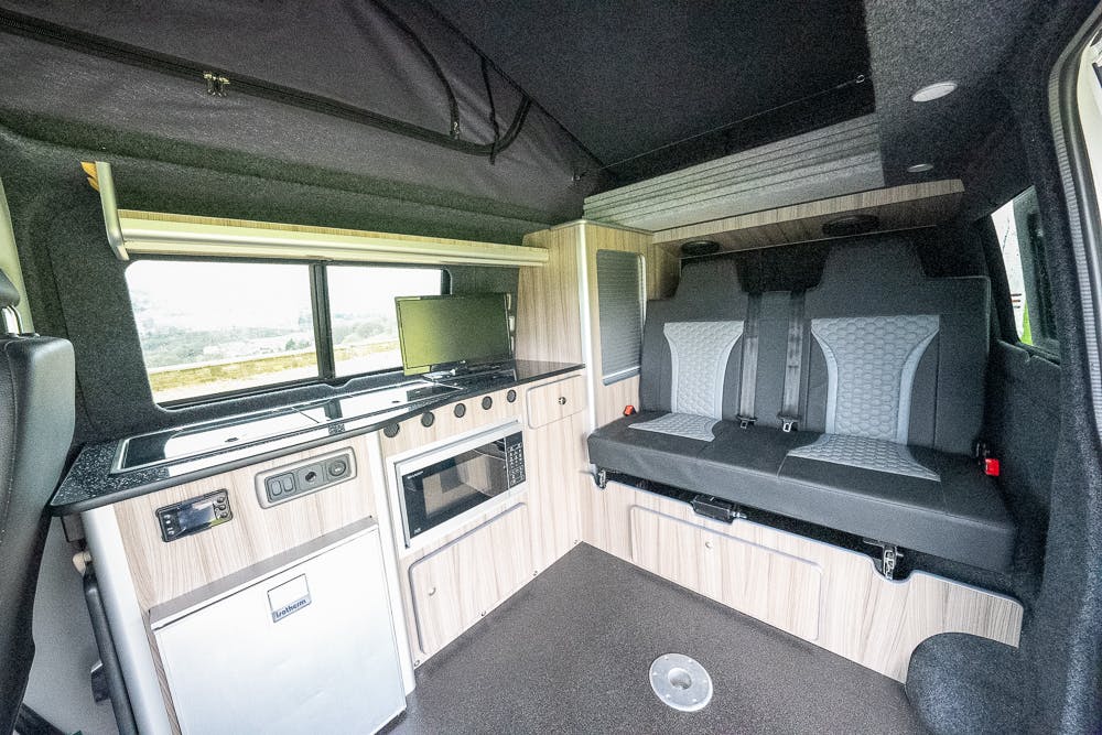 The interior of the 2019 Volkswagen Transporter T28 Trendline TDI camper van includes a compact kitchen with a small refrigerator, microwave, stovetop, and sink. Adjacent to the kitchen is a seating area with seat belts, upholstered in gray and black. A window provides natural light in the space.