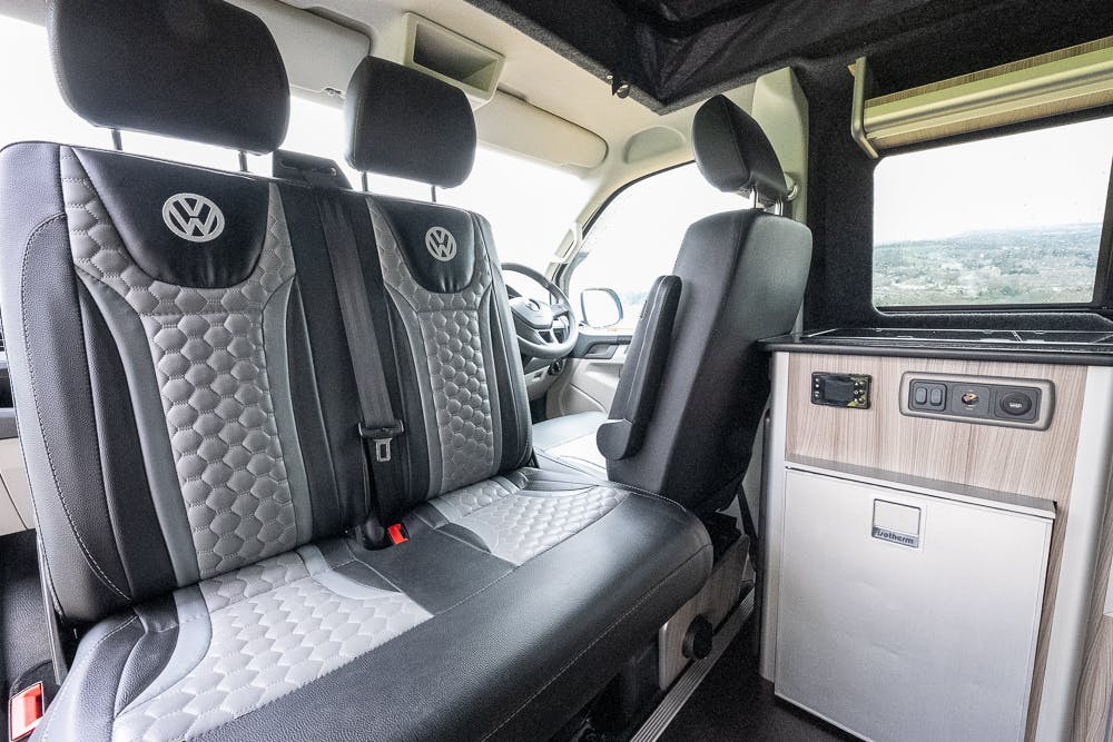 Interior of a 2019 Volkswagen Transporter T28 Trendline TDI, featuring two-tone black and grey leather seats with a Volkswagen logo, a compact kitchen area with a small refrigerator, storage compartments, and a countertop. The driver's seat is partially visible.