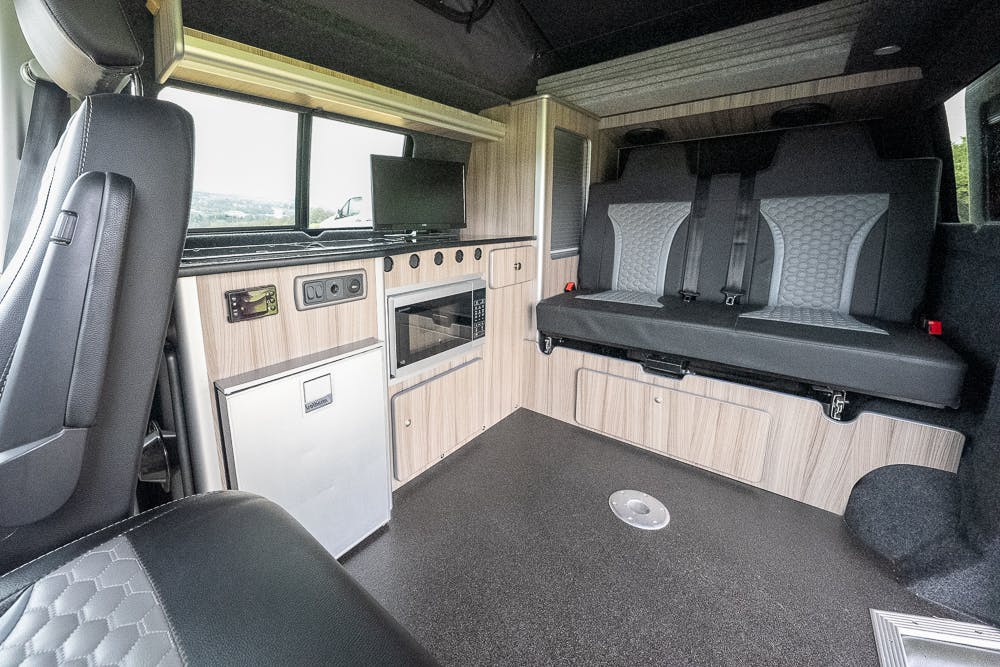 The interior of the 2019 Volkswagen Transporter T28 Trendline TDI features a compact kitchenette with a microwave, mini refrigerator, and overhead storage cabinets. Adjacent is a bench seat with seatbelts, and the flooring is dark with a round drain in the center. A window provides outside light.