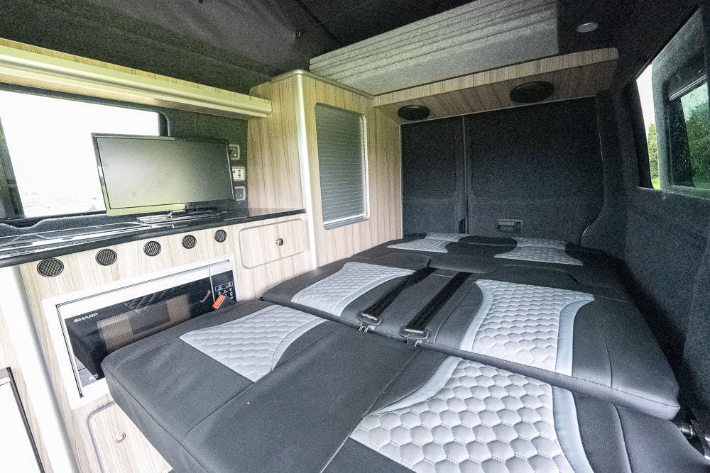 The image shows the interior of a 2019 Volkswagen Transporter T28 Trendline TDI camper van with a bed setup. The space includes a flat-screen TV mounted on the left wall, a kitchen area with a built-in microwave, and overhead storage cabinets. The bed has a grey cushioned surface and black accents.