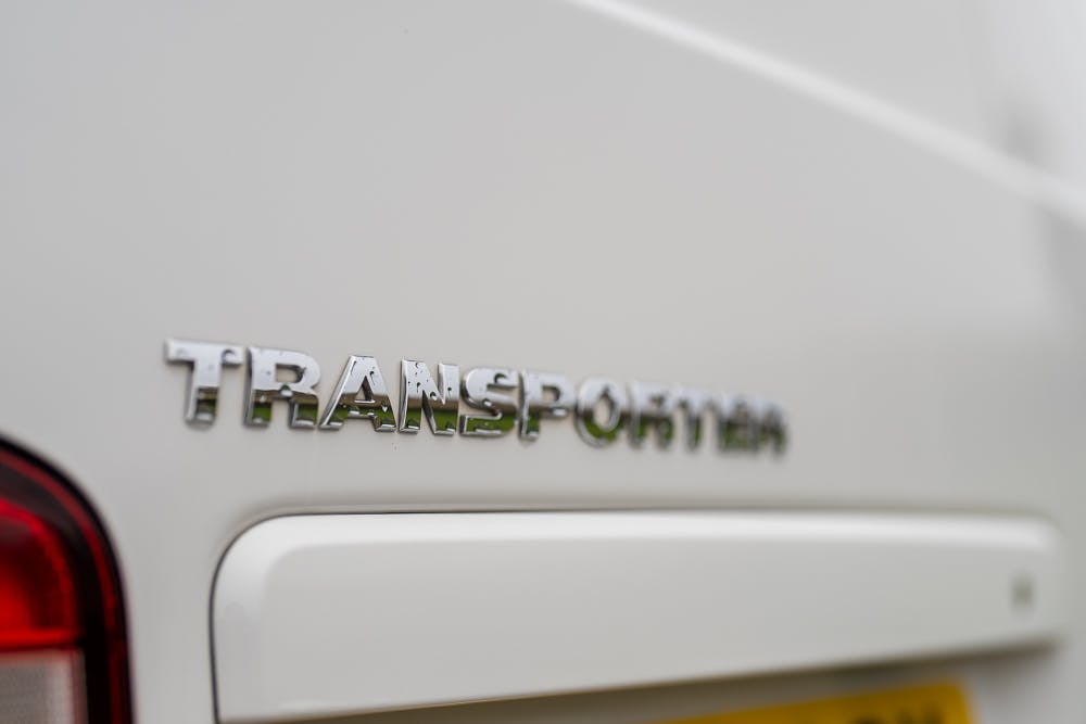 Close-up image of the rear of a 2019 Volkswagen Transporter T28 Trendline TDI in white, with the word "Transporter" in chrome letters. The shot includes a partial view of a red light on the left and a yellow license plate at the bottom right.