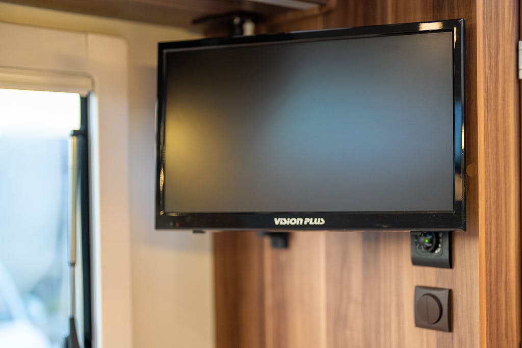 A flat-screen television with the brand name "Vision Plus" is mounted on a wooden wall inside a 2016 Roller Team Auto-Roller 707 Low Line. Below the TV, there is a black electrical switch and an integrated webcam. A portion of a window is visible to the left.