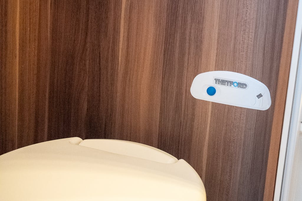 A close-up image of a Thetford toilet in a restroom with wood panel walls, located inside a sleek 2016 Roller Team Auto-Roller 707 Low Line. The Thetford brand name is visible on a small control panel mounted on the wall above the toilet, featuring a blue button.