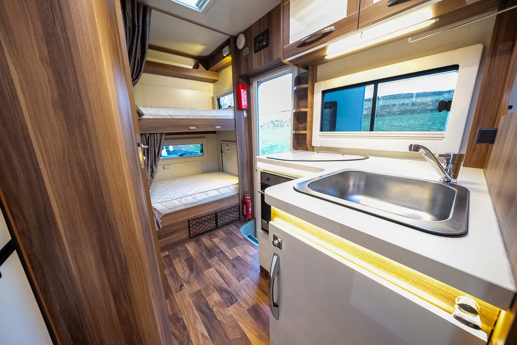 Interior of the 2016 Roller Team Auto-Roller 707 Low Line camper van featuring a small kitchen with a sink, countertop, storage cabinets, and a mini fridge. The adjacent area includes a sleeping space with bunk beds and wooden flooring throughout. A window provides natural light.