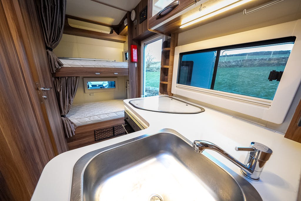 The interior of the 2016 Roller Team Auto-Roller 707 Low Line features a compact RV design with a small kitchen, complete with a stainless steel sink and faucet in the foreground. To the left, cozy bunk beds with privacy curtains are situated, and above the counter, a window offers a view of green fields.