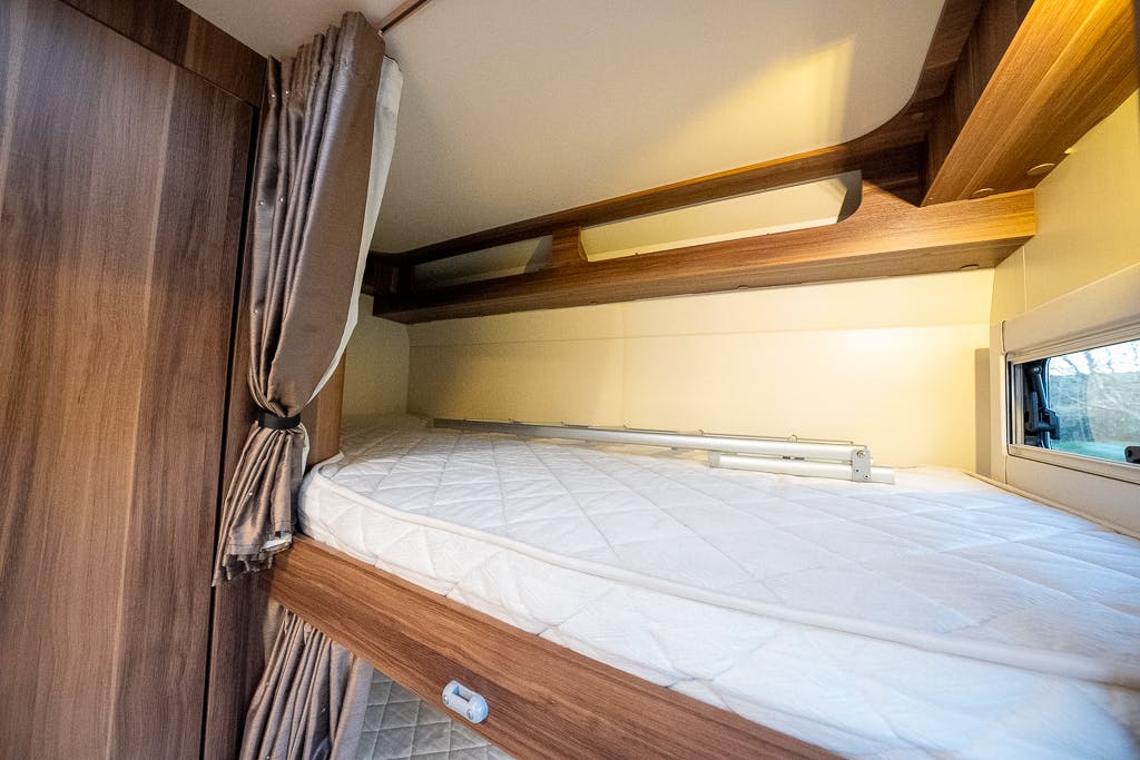The image shows a neatly made upper bunk bed inside a 2016 Roller Team Auto-Roller 707 Low Line. The bed has a white mattress with a quilted cover, surrounded by wooden panels. A small rectangular window with a white blind is visible on the right side.