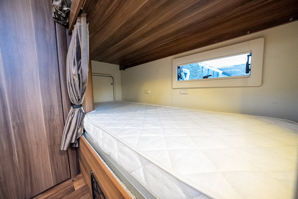 A small bunk bed with a white mattress and brown curtains on the side is neatly integrated into the 2016 Roller Team Auto-Roller 707 Low Line. The wooden-framed bed has a horizontal window above it, showcasing the exterior view. The interior boasts a wooden finish and minimal decor, perfect for cozy travels.