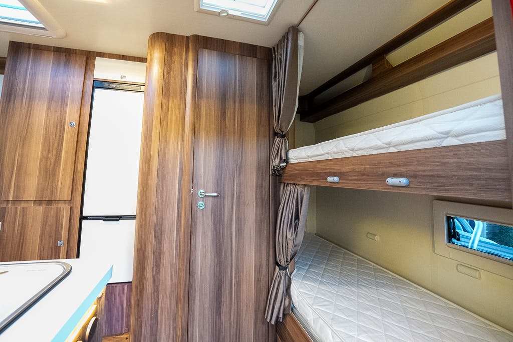 The interior of the 2016 Roller Team Auto-Roller 707 Low Line camper van features wooden paneling, a small window, and a set of bunk beds with white mattresses and privacy curtains. The kitchenette is visible on the left side, and a closed door is in the center.