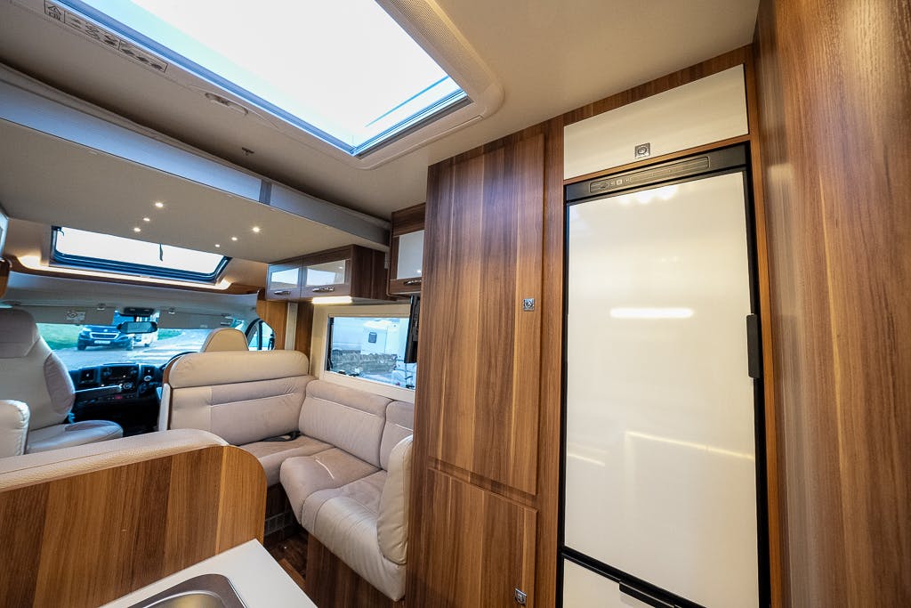 Interior of the 2016 Roller Team Auto-Roller 707 Low Line featuring a skylight, cream-colored sofa seating, and wooden cabinetry. A white refrigerator is visible on the right, and a sink is partially visible in the foreground. The motorhome has large windows and ceiling lights.
