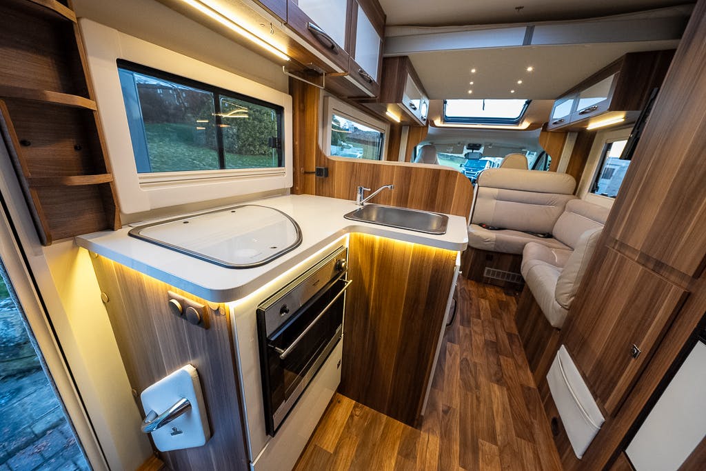 The image shows the interior of a 2016 Roller Team Auto-Roller 707 Low Line motorhome featuring a kitchen area with a sink and stovetop, a small oven, wooden cabinets, and drawers. Adjacent to the kitchen is a seating area with white leather upholstery and windows allowing natural light inside.