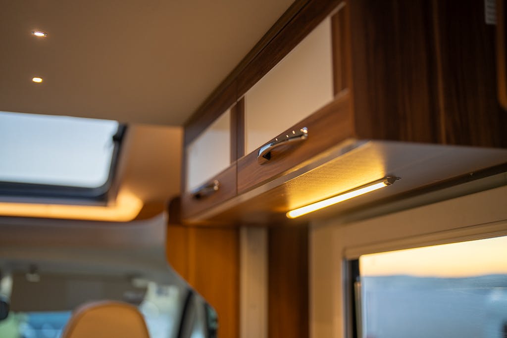 The image shows the interior of a 2016 Roller Team Auto-Roller 707 Low Line, featuring wooden overhead cabinets with silver handles. Below the cabinets, a strip light is illuminated. To the right, a window is partially visible. The ceiling includes a skylight, emitting natural light into the space.