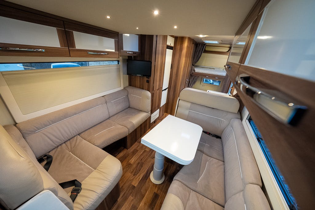 Interior of a 2016 Roller Team Auto-Roller 707 Low Line motorhome with wood-paneled walls, beige cushioned seating, and a white rectangular table in the center. Overhead cabinets provide storage, and a small TV is mounted on the wall. The space is brightly lit with recessed lighting.
