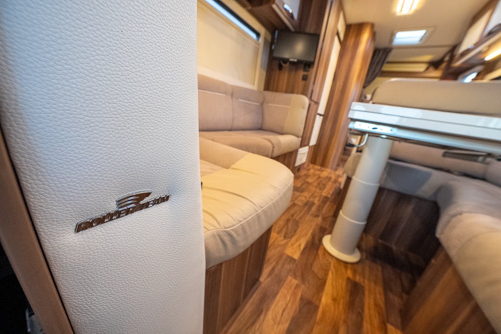 The image shows the interior of a 2016 Roller Team Auto-Roller 707 Low Line luxury motorhome. There are beige cushioned seating areas, wooden flooring, a table with a central support pole, and a mounted flat-screen TV. The corner of a seating area features a metallic emblem.