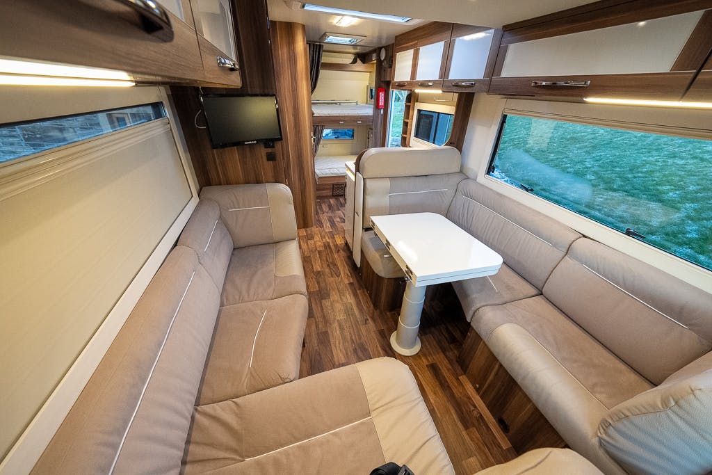 Interior of the 2016 Roller Team Auto-Roller 707 Low Line features a seating area with beige cushions surrounding a white table, wooden cabinetry, a small wall-mounted TV, and a hallway leading to bunks in the back. Large windows on both sides provide natural light.