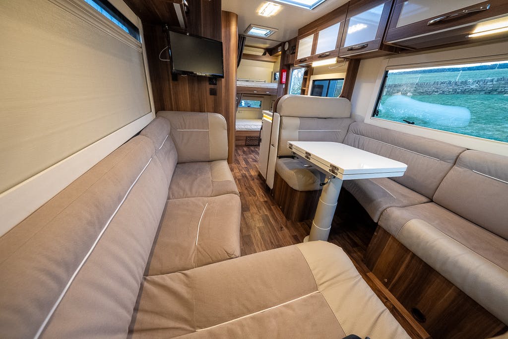 Interior of the 2016 Roller Team Auto-Roller 707 Low Line camper van featuring a beige and brown color scheme. The space includes two large sofas, a foldable dining table, a wall-mounted TV, various overhead storage cabinets, and a view of the outdoors through the windows.