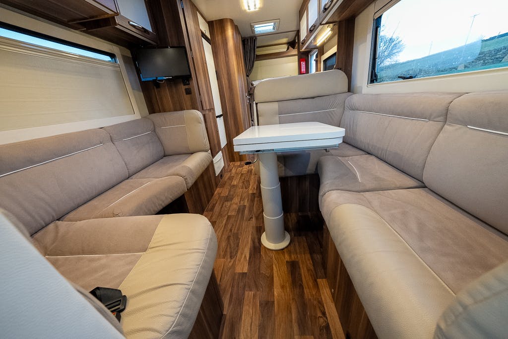 The interior of the 2016 Roller Team Auto-Roller 707 Low Line motorhome features tan cushioned seating along both walls. A white table sits in the center on a single pedestal. Wooden flooring and cabinets are visible. A window with a roller shade is on the left, and a small TV is mounted on the wall.