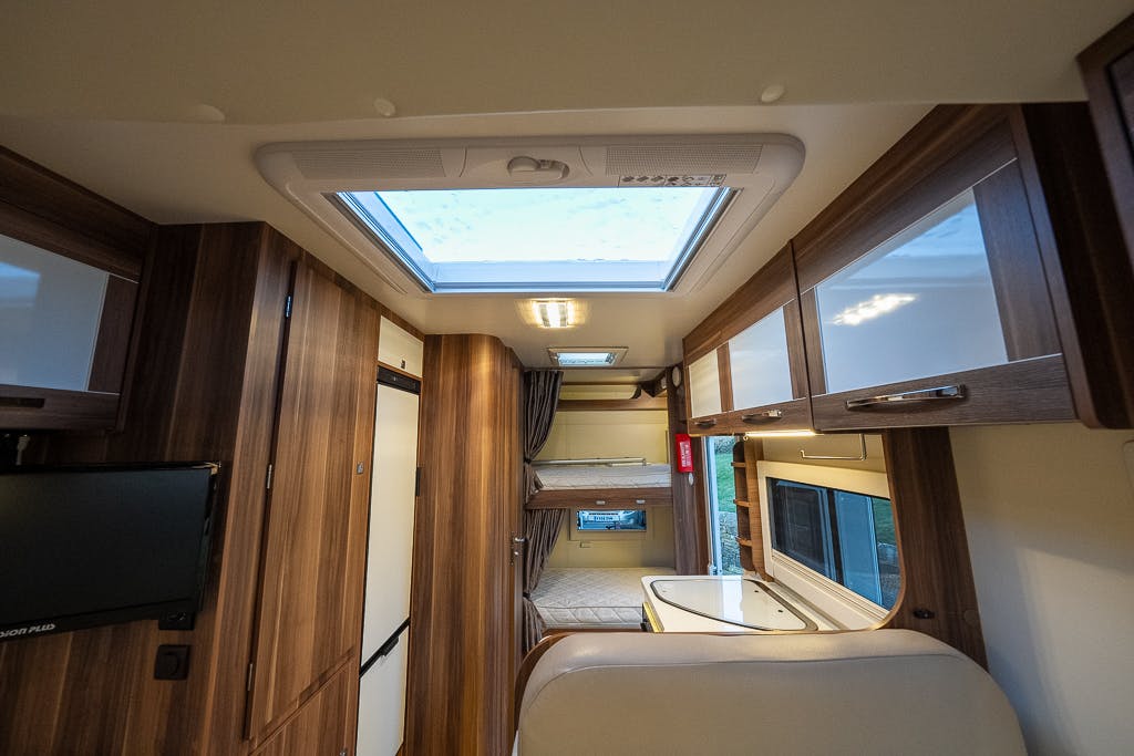 The interior of the 2016 Roller Team Auto-Roller 707 Low Line RV features wood-paneled cabinets, a skylight, a flat-screen TV, and a dining area. The RV has a clean and organized layout with a passageway leading to a rear sleeping area.