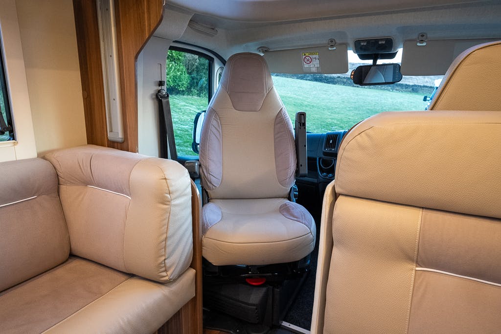 The interior of the 2016 Roller Team Auto-Roller 707 Low Line motorhome features beige seating, with a driver's seat in the center facing backwards. The living area includes a cushioned bench on the left side. Green grass is visible through the windshield.