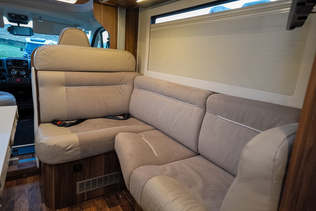 This image shows the interior of a 2016 Roller Team Auto-Roller 707 Low Line camper van with beige upholstered seating arranged in an L-shape. The seating area is next to a wall with a large window. The floor is made of wood, and the driver’s seat is partially visible.