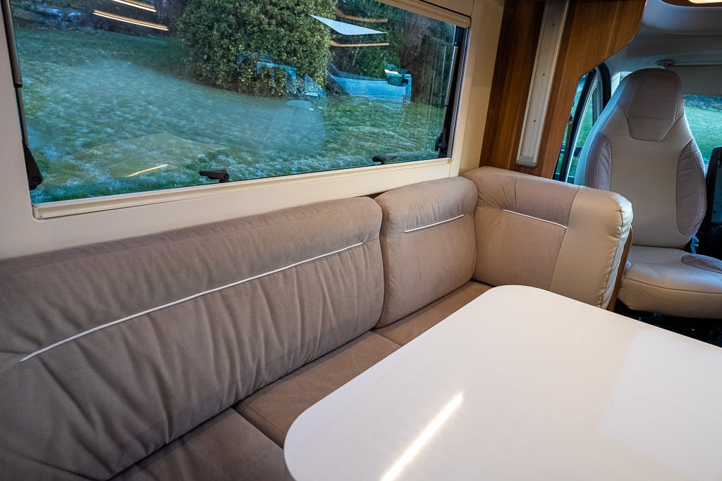 The interior of the 2016 Roller Team Auto-Roller 707 Low Line motorhome is shown with a beige cushioned sofa along the side wall, a white table in front of it, and a beige driver's seat partially visible. The window offers a view of a grassy area outside.