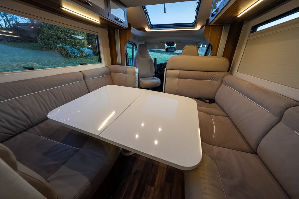 The interior of the 2016 Roller Team Auto-Roller 707 Low Line motorhome is shown with a beige and gray color scheme. There is a U-shaped seating arrangement with a white table in the center. The area is well-lit with natural light coming through the windows and a skylight.