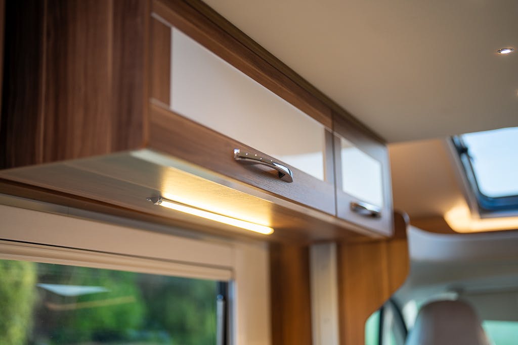 Interior view of a wood-paneled overhead cabinet in the 2016 Roller Team Auto-Roller 707 Low Line. The cabinet has metallic handles and is illuminated by a strip light below. A window and partially open skylight can be seen in the background.