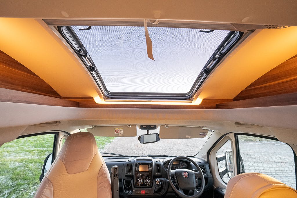 The image shows the front seating area of a 2016 Roller Team Auto-Roller 707 Low Line camper van. The picture is taken from the middle of the van, showcasing two seats, a steering wheel, and various vehicle controls. A large sunroof with wooden trim is visible above the seats.