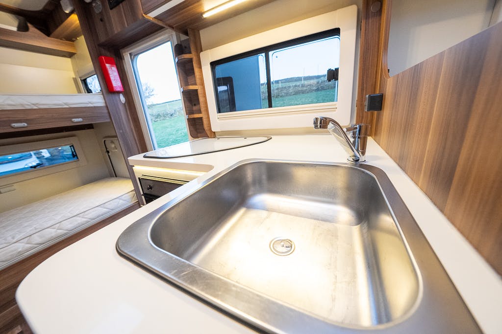 The image shows the interior of a 2016 Roller Team Auto-Roller 707 Low Line, focusing on the sink area. The stainless steel sink is installed on a white countertop with a faucet. In the background, there is a bunk bed and a window bringing in natural light. The walls have a wooden finish.