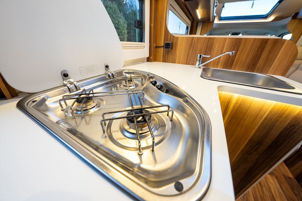 A modern campervan kitchen in the 2016 Roller Team Auto-Roller 707 Low Line features a stainless steel three-burner gas stove and a matching sink. The white countertop contrasts beautifully with the wooden cabinetry, while a window offers a picturesque view of greenery.