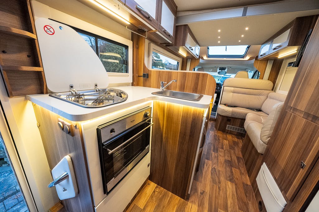 The image shows the interior of a 2016 Roller Team Auto-Roller 707 Low Line. It features a small kitchen area with a stove, oven, and sink on a wooden countertop. There is a white couch in the background, wood-paneled cabinets, and a skylight with recessed lighting. The floor is wood-patterned.