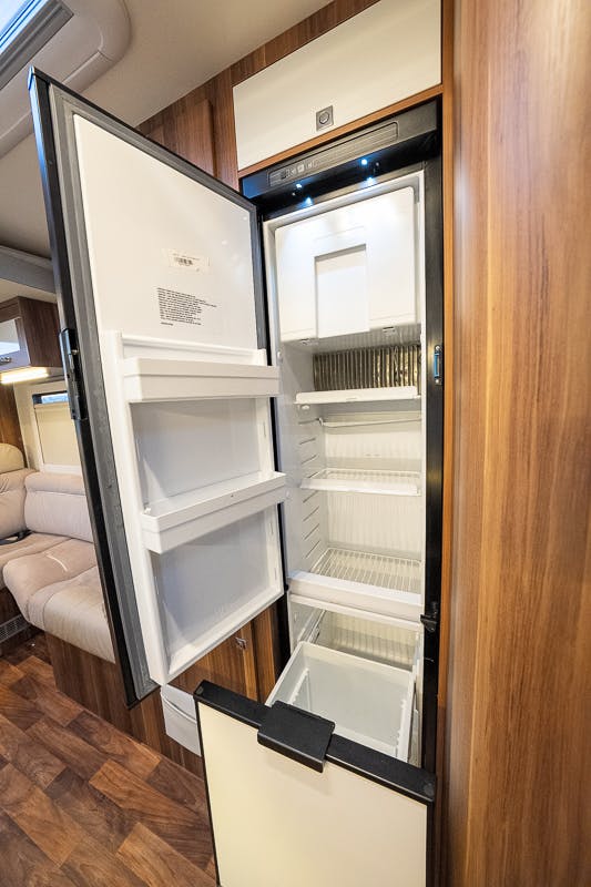 An open, empty refrigerator is situated in the cozy wooden kitchenette area of the 2016 Roller Team Auto-Roller 707 Low Line. Built into a cabinet, the fridge features a top freezer compartment and a main refrigerator section below. The surrounding area offers comfortable beige cushioned seating.
