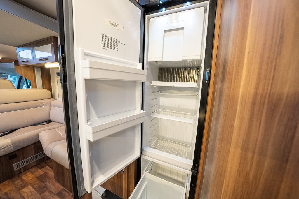 An open refrigerator in a 2016 Roller Team Auto-Roller 707 Low Line motorhome's kitchen area. The fridge has several empty shelves and door compartments. In the background, there is a beige seating area with cushioned seats, wood paneling, and part of the motorhome's interior visible.