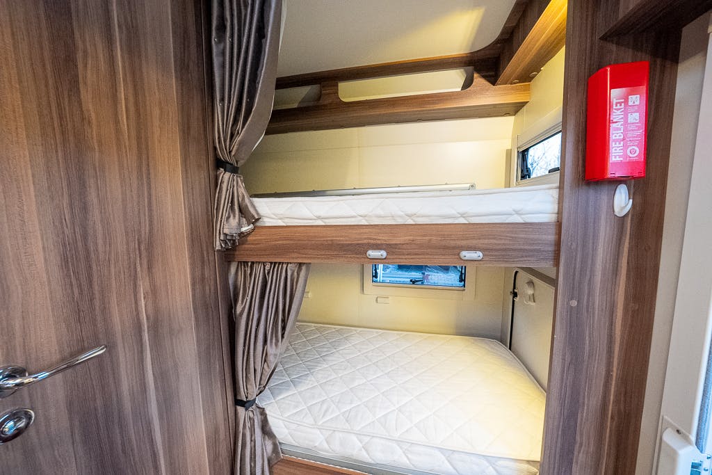 The image shows a set of bunk beds in a small room, likely within a 2016 Roller Team Auto-Roller 707 Low Line RV. The mattresses are bare, and a curtain is used to provide privacy for the lower bunk. A red fire blanket is mounted on the wooden wall next to the bunks.